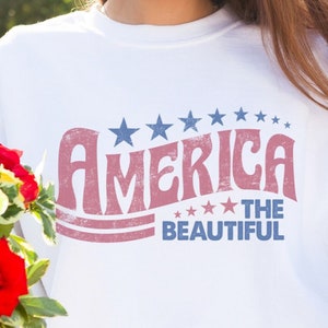 May include: White sweatshirt with the text "America The Beautiful" in red and blue. The design includes stars above and below the text. The sweatshirt is worn with blue jeans.