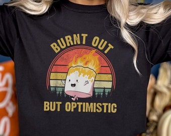 May include: Black t-shirt featuring a cartoon marshmallow on fire with the text "BURNT OUT BUT OPTIMISTIC". The design is set against a sunset background. The person is wearing a beige beanie and blue jeans.
