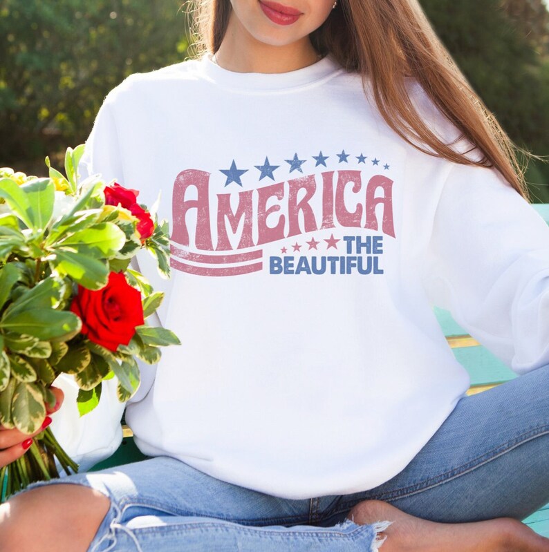 May include: White sweatshirt with the text "America The Beautiful" in red and blue. The design includes stars above and below the text. The sweatshirt is worn with blue jeans.