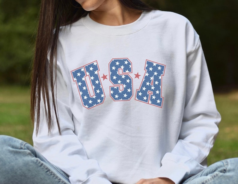 May include: White long-sleeve sweatshirt with the word "USA" in blue letters filled with white stars and outlined in red. Two red stars are on either side of the letters. The sweatshirt is a casual style.
