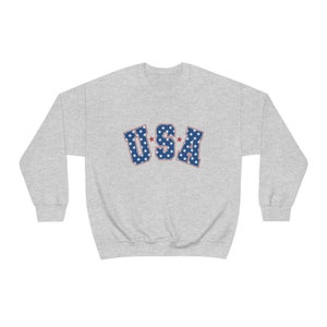 May include: A light gray sweatshirt with the word "USA" in blue letters filled with white stars and outlined in red. The sweatshirt is a crew neck style.