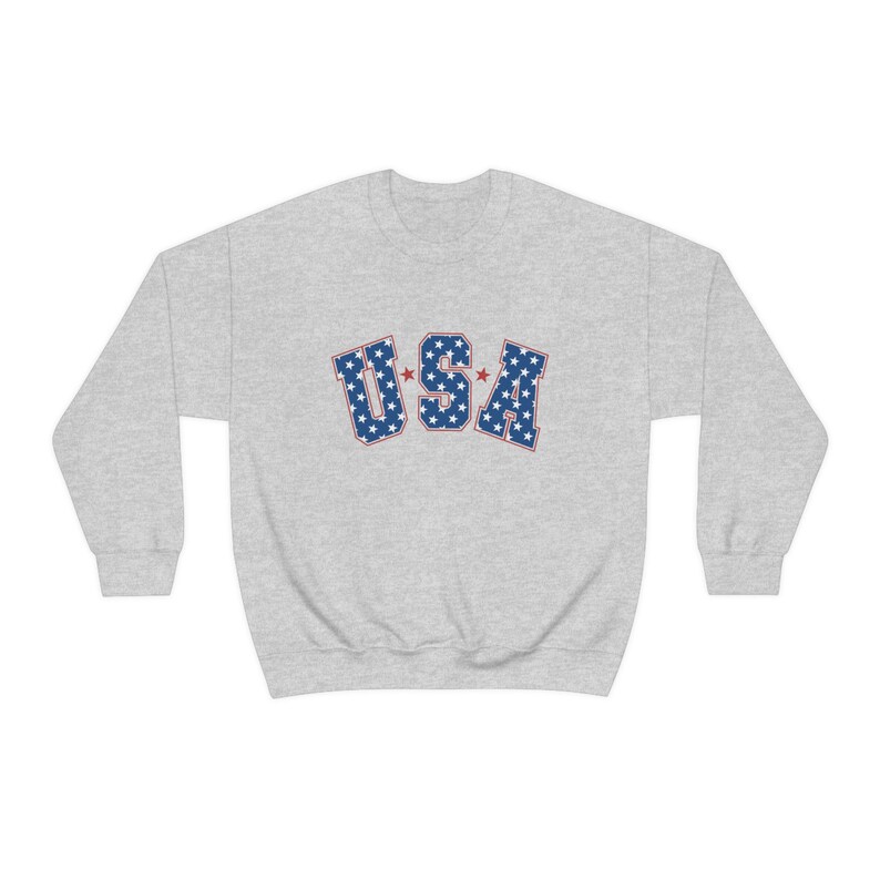 May include: A light gray sweatshirt with the word "USA" in blue letters filled with white stars and outlined in red. The sweatshirt is a crew neck style.