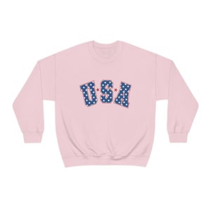 May include: A light pink sweatshirt with the word "USA" in blue letters filled with white stars. The letters have a red outline. The sweatshirt has long sleeves and a crew neck.