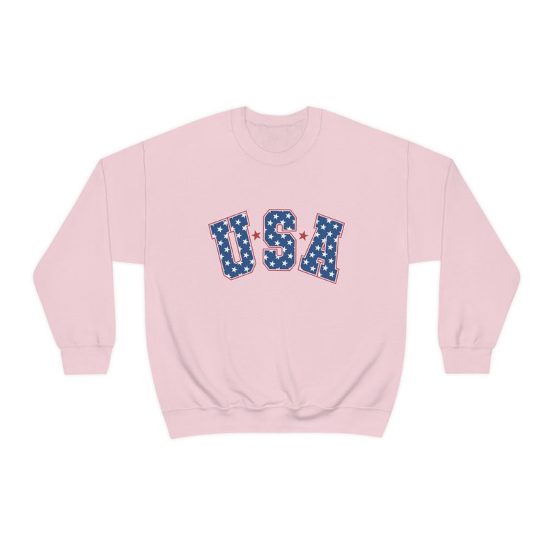 May include: A light pink sweatshirt with the word "USA" in blue letters filled with white stars. The letters have a red outline. The sweatshirt has long sleeves and a crew neck.