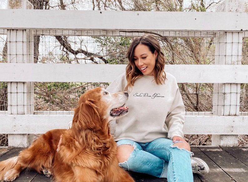 May include: A woman wearing a beige sweatshirt with the text "Soli Deo Gloria" in black script, sits on a wooden deck with a golden retriever dog. The woman is smiling and looking at the dog.