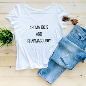 May include: White v-neck t-shirt with the text "Aroma Joe's and Pharmacology" printed on the front. The shirt is laid out on a wooden surface with a pair of blue jeans and sunglasses.