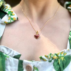 Puede incluir: Un collar dorado con un colgante en forma de corazón. El colgante es rojo con un borde dorado. La persona lleva un vestido blanco con estampados de hojas verdes y amarillas.