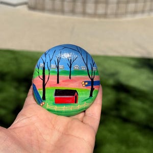 May include: Hand-painted rock art depicting a rural scene with a red house, trees, and a blue sky. The rock is painted in a folk art style.
