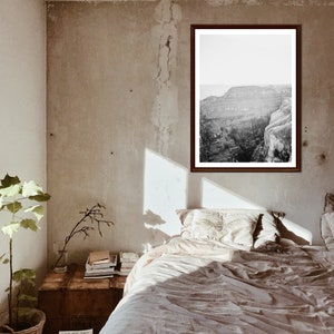 Puede incluir: Una fotografía en blanco y negro de un paisaje de cañón está enmarcada y colgada en una pared sobre una cama con ropa de cama blanca. La pared es de color beige claro con una textura rugosa.