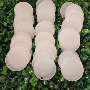 Op de afbeelding: Een stapel witte, ronde, blanco acryl cirkels met een gat in het midden. De cirkels zijn gerangschikt in rijen op een groen, bladerrijk oppervlak.