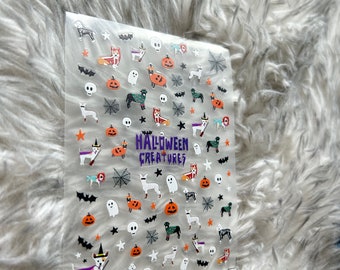 May include: A sheet of clear nail stickers with a Halloween theme. The stickers feature various images, including pumpkins, ghosts, bats, stars, skulls, spiderwebs, and dogs dressed in Halloween costumes. The text "HALLOWEEN CREATURES" is printed in purple on the sheet.