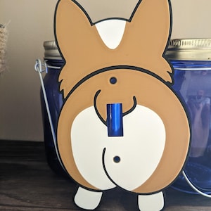 May include: A novelty light switch cover in the shape of a corgi's rear end. The cover is brown, white, and black, with a blue switch. The ears are upright, and the design includes a smiling expression. The cover is mounted on a wooden surface.