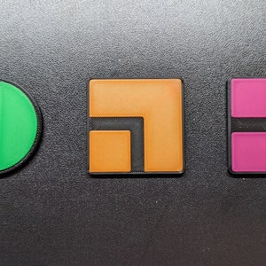 May include: Three colorful geometric shapes: a green circle with a black stripe, an orange square with an L-shape cut out, and a pink square with four smaller squares. All shapes have a black border.