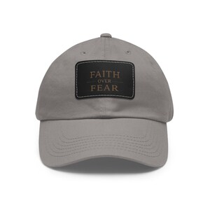 May include: A gray baseball cap with a curved brim. The cap features a rectangular black patch with the words "FAITH OVER FEAR" in gold lettering. The hat is made of a soft material and has a classic, casual style.