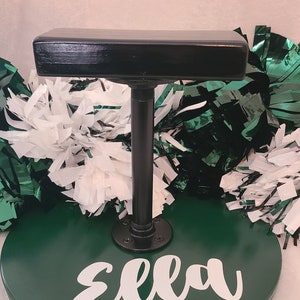 Op de afbeelding: Een groen en zwart cheerleading-rekwisiet met de naam "Ella" in wit script. Het rekwisiet heeft een ronde basis, een zwarte metalen paal en een zwart rechthoekig bovenstuk. Witte en groene pompons staan op de achtergrond.