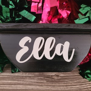 May include: A black wooden shelf with the name "Ella" in white script. The shelf has a curved front and a flat top. Green and pink metallic fringe is in the background.