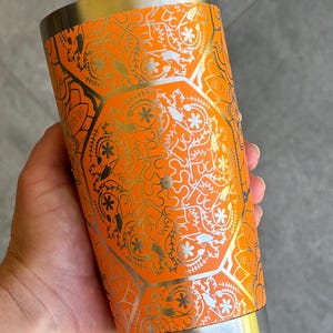 May include: Orange stainless steel tumbler with a gold metallic design featuring a repeating pattern of flowers, leaves, and geometric shapes.