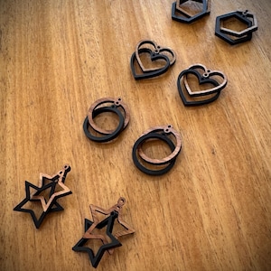 May include: A set of wooden earrings in various shapes, including hearts, stars and hexagons. The earrings are made of two layers of wood, with the top layer being black and the bottom layer being a natural wood colour.