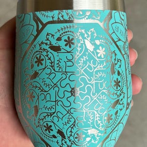 Puede incluir: Un vaso de acero inoxidable turquesa con un dise&ntilde;o met&aacute;lico plateado que presenta un patr&oacute;n repetitivo de flores, hojas e insectos. El texto "F*CK OFF" es visible en el lateral del vaso.