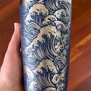 May include: A stainless steel tumbler with a navy blue background and a silver wave pattern. The design features stylized ocean waves. The tumbler has a silver rim at the top and bottom. The tumbler is approximately 18 cm tall.