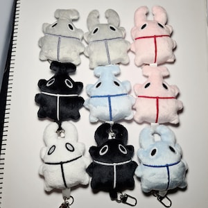 May include: A collection of nine plush beetle keychains arranged in a three-by-three grid. The soft toys feature embroidered eyes and a central vertical line detail. They come in various colours including white, black, light blue, and pale pink.