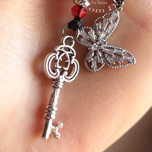 May include: A silver key charm with an ornate design hangs from a red and white beaded bracelet. The bracelet also features a silver butterfly charm with clear rhinestones. The bracelet is labelled "Blythes Little Custom Dolls by Tanya".
