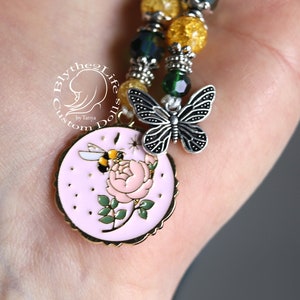 May include: A pink charm with a bee and a rose, a silver butterfly charm, and a yellow and green beaded necklace. The charm is attached to the necklace.