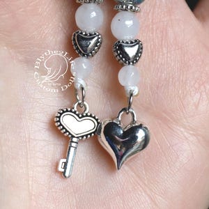 May include: A white and silver charm bracelet with a key charm, a heart charm, and a heart-shaped bead. The bracelet is made of white beads and silver metal.