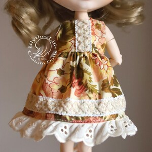 May include: A doll wearing a floral print dress with lace trim and a white lace petticoat. The dress has a brown, yellow, and orange floral pattern. The doll has blonde hair and blue eyes. The doll is wearing a dress that says "Blythe the Life Custom Dolls by Tanya".