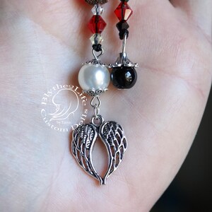 May include: A silver charm with angel wings hangs from a beaded necklace with black and red beads. The necklace also features a white pearl and a black bead.