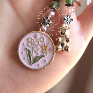 May include: A pink and gold charm with a lily flower and butterfly design. The charm is attached to a green and brown beaded necklace with a silver bear charm.