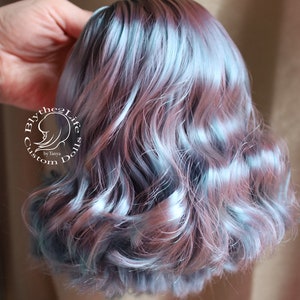 May include: A doll wig with wavy, shoulder-length hair. The hair is a blend of blue, pink, and gray tones, creating a soft, pastel effect. The wig is styled with gentle curls and has a natural appearance. The text "Blythe2Life Custom Dolls by Tanya" is visible.