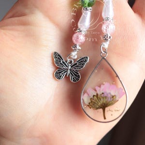 May include: A silver butterfly charm hangs from a beaded necklace with pink and white beads. A teardrop-shaped pendant with a pressed pink flower inside is attached to the necklace.