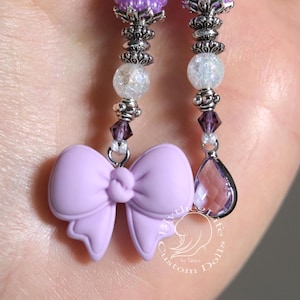 May include: A pair of purple and silver earrings with a lavender bow charm and a teardrop-shaped crystal. The earrings are made with beads and have a silver metal base.