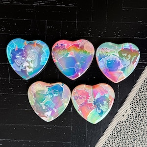 Puede incluir: Cinco botones iridiscentes en forma de corazón con ilustraciones coloridas de personajes de dibujos animados. Cada botón presenta un par diferente de personajes en varias poses. Los botones están dispuestos sobre una superficie oscura.