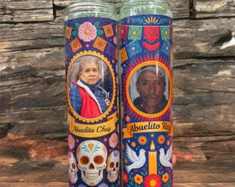 May include: Two glass jar candles with portraits and decorative designs. One candle features a woman's portrait with the text "Abuelita Chuy" and sugar skull imagery. The other candle has a man's portrait with the text "Abuelito Rey" and dove imagery.