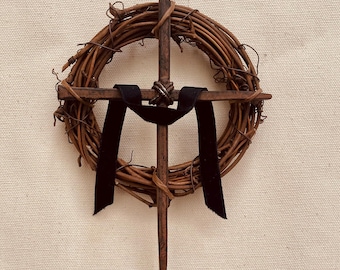 May include: A rustic wooden wreath with a metal cross in the center. The cross is made of a single piece of metal and has a black ribbon tied around it. The wreath is made of brown twigs and is about 12 inches in diameter.