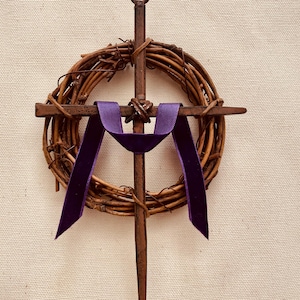 May include: A rustic wooden cross with a purple ribbon tied around it. The cross is decorated with a wreath of brown twigs.