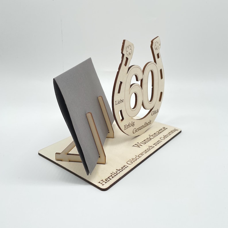 May include: Wooden card holder with a horseshoe design and the number 60 in the center. The horseshoe has the words "Liebe", "Erfolg", "Gesundheit", and "Gl&uuml;ck" inscribed around it. The base of the holder has the text "Herzlichen Gl&uuml;ckwunsch zum Geburtstag" and "W&uuml;nsche" inscribed on it.