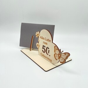 May include: Wooden card holder with a heart-shaped design. The heart has a butterfly and the text "Alles Liebe zum 50. Geburtstag".