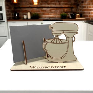 Könnte beinhalten: Holzständer mit einem Ausschnitt eines Küchenroboters und einem grauen Kartenhalter. Der Ständer hat den Text "Wunschtext" auf der Basis graviert.