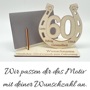 May include: Wooden horseshoe-shaped birthday card holder with the number 60 in the center. The horseshoe has the words "Liebe", "Erfolg", "Gesundheit", and "Gl&uuml;ck" inscribed on it. The card holder has a small stand to hold a card. The text "Herzlichen Gl&uuml;ckwunsch zum Geburtstag" is inscribed on the base of the stand. The text "Wir passen dir das Motiv mit deiner Wunschzahl an." is written below the image.