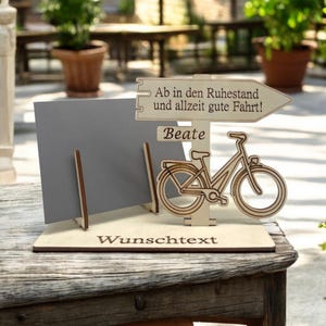 May include: A wooden sign with a bicycle cutout, a gray card, and text. The sign reads "Ab in den Ruhestand und allzeit gute Fahrt!" and "Beate". The base has the word "Wunschtext". The sign is on a wooden table.