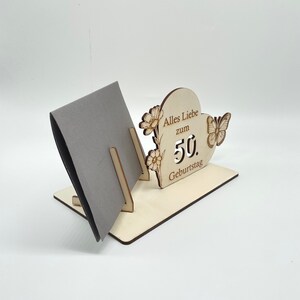 May include: Wooden card holder with a heart-shaped cutout and the text "Alles Liebe zum 50. Geburtstag". The holder has a gray card inside and a butterfly design.