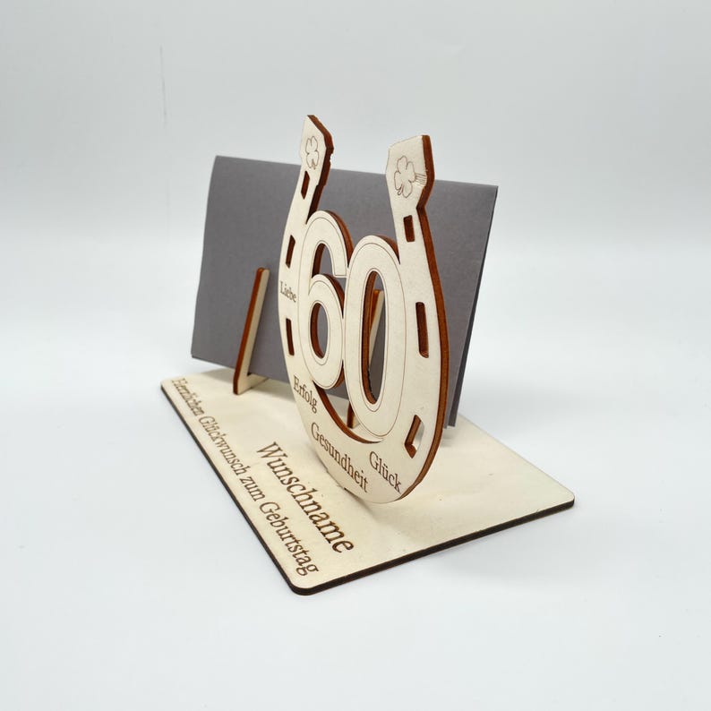 May include: Wooden card holder shaped like a horseshoe with the number 60 in the center. The holder has a slot for a card and a stand to hold it upright. The words "Wunschname", "Gl&uuml;ck", "Gesundheit", "Erfolg", and "Liebe" are engraved around the horseshoe. The words "Herzlichen Gl&uuml;ckwunsch zum Geburtstag" are engraved on the base.