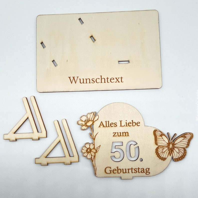 May include: Wooden craft kit for a 50th birthday. The kit includes a heart-shaped plaque with the text "Alles Liebe zum 50. Geburtstag" and a butterfly, two wooden stands, and a blank plaque for a personalized message.