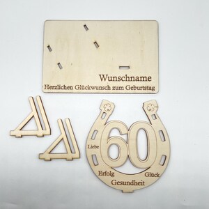 May include: Wooden craft kit for a 60th birthday celebration. The kit includes a horseshoe with the number 60 inside, two small triangles, and a sign that says "Wunschname Herzlichen Gl&uuml;ckwunsch zum Geburtstag". The horseshoe has the words "Liebe", "Erfolg", "Gesundheit", and "Gl&uuml;ck" around it.