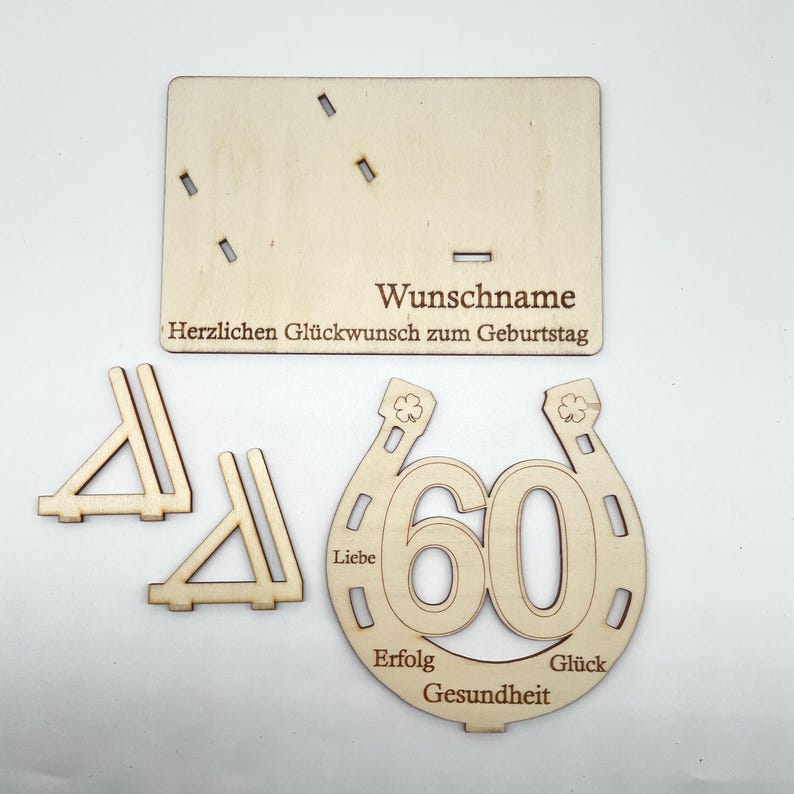 May include: Wooden craft kit for a 60th birthday celebration. The kit includes a horseshoe with the number 60 inside, two small triangles, and a sign that says "Wunschname Herzlichen Gl&uuml;ckwunsch zum Geburtstag". The horseshoe has the words "Liebe", "Erfolg", "Gesundheit", and "Gl&uuml;ck" around it.