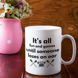 May include: White ceramic mug with the text "It's all fun and games until someone loses an oar." The mug features a crab and crossed oars graphic. A pink vase with white polka dots and a bouquet of flowers is in the background.