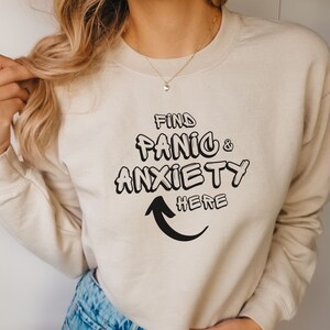 May include: A beige sweatshirt with the text "Find Panic & Anxiety Here" in black, with an arrow pointing down.
